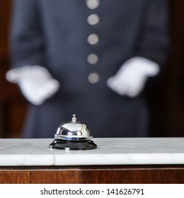 Concierge With White Gloves Pointing To Hotel Bell On Counter