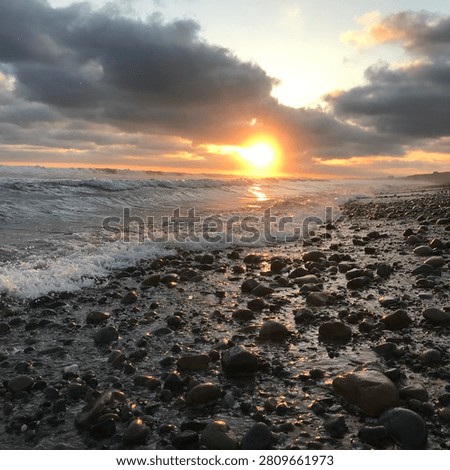 Concise: A beautiful photograph capturing a dramatic sunset over a rocky coastline with waves crashing onto the pebble shore.