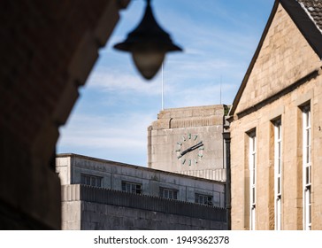 Concrete Art Deco Clock Tower With Roman Numerals On Top Of Retro Department Store In Town Centre Viewed From Under Bridge Archway With 1950's Old Out Of Focus Street Lamp Hanging Down.