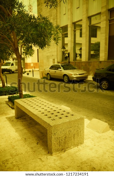 Concrete bench of Santo Domingo street. Bench is\
lit with soft sun light falling through the clouds in warm autumn\
day. Bench stands on sidewalk. Some old cars are parked near.\
Vintage style photo