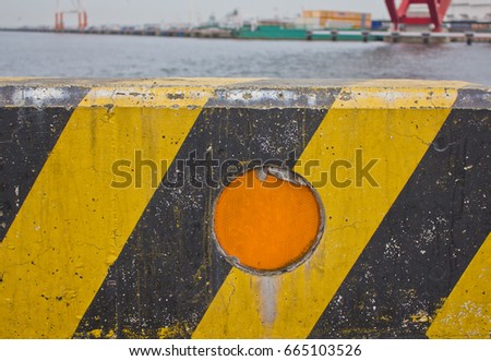A concrete block painted with a yellow and black zigzag pattern and featuring a circular orange safety reflector marks the end of the road at a harbor in Kobe, Japan.
