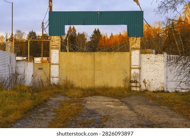 Concrete and brick security gate blocking access to abandoned industrial lot, rusted metal fencing topped with barbed wire, cracked asphalt driveway with grass and