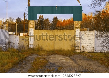 Concrete and brick security gate blocking access to abandoned industrial lot, rusted metal fencing topped with barbed wire, cracked asphalt driveway with grass and