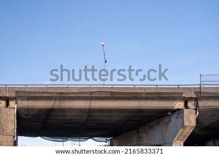 A concrete bridge over a two lane highway. The high overpass has a metal handrail. Black construction netting hangs from under the bridge to catch any concrete debris during construction repairs.