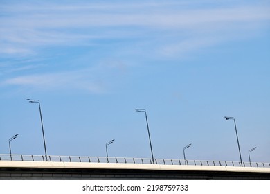 Concrete City Bridge Over River With Street Lampposts, Angle View With Blue Sky Copy Space Background. Minimalistic Bridge With Automobile Highway And Wireless Lamp Posts, Modern City Infrastructure