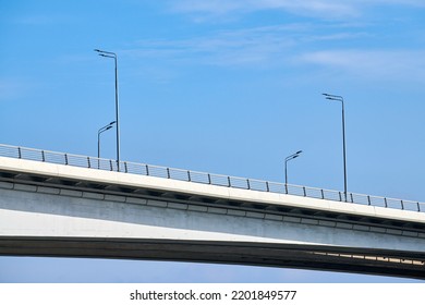 Concrete City Bridge Over River With Street Lampposts, Blue Sky Background. Minimalistic Bridge With Automobile Highway And Wireless Lamp Posts, Modern City Infrastructure