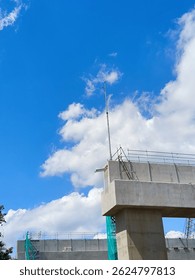 Concrete column structure of an elevated toll road under construction, with temporary scaffolding and weather monitoring equipment installed on top. Clear blue sky in the background.