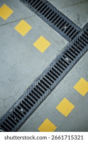 Concrete floor featuring intersecting metal drainage grates and diagonal rows of yellow square markings, geometric industrial pattern emphasizing urban infrastructure elements
