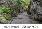 A concrete footpath runs between the rocks. Green vegetation on weathered, cracked slopes. Puddles of water on a wet path. China. Tianxingqiao Scenic Area. Heavenly Star Bridge Tianxing Qiao.