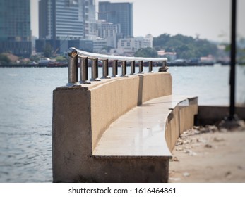 Concrete Long Bench, Stainless Steel Railing On Top, Located On The Water Front, Coastal Town As A Backdrop.