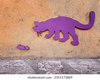 a concrete orange wall with purple wooden cat stalking a small mouse , funny purple cat chasing a scaredy small mouse in a cement wall, street decoration, horizontal