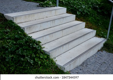 Concrete Stairs To The Park Serving At The Same Time As A Sitting Bench. Gray Cement Clean Smooth Bright Cement Surface. Granite Cubes Paving Threshing Gravel Road. Overgrown With Plants. Anti-slip