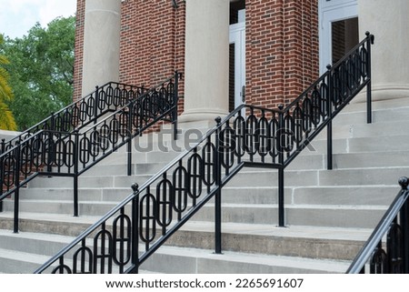 Concrete steps lead to multiple doors of a historic building. The walls of the building are made of red brick. There are black decorative metal handrails dividing the stairs to the entrance.