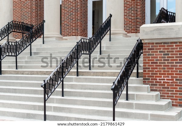 Concrete steps lead to multiple doors of a\
historic building. The walls of the building are made of red brick.\
There are black decorative metal handrails dividing the stairs to\
the entrance.