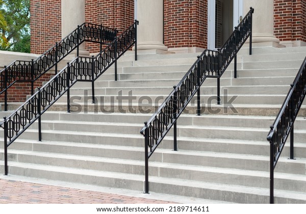 Concrete steps lead to multiple doors of a\
historic building. The walls of the building are made of red brick.\
There are black decorative metal handrails dividing the stairs to\
the entrance.