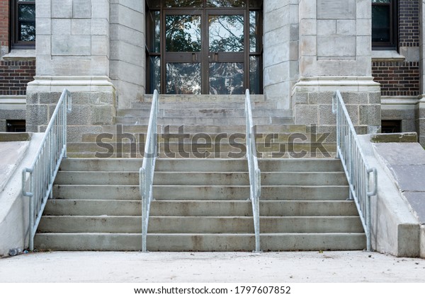 Concrete steps lead to a red double door of a\
historic building. The wall of the building is made of light grey\
granite block.There are four metal handrails dividing the stairs to\
the entrance.