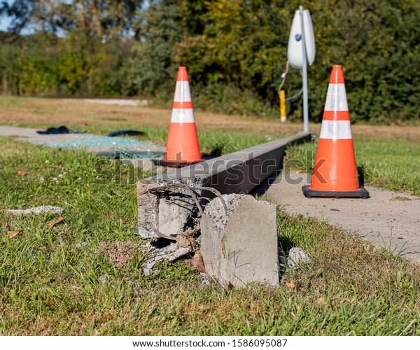 Concrete streetlight pole broken from car\
accident, with exposed wires and laying across sidewalk. Orange\
traffic cones and shattered glass in\
background