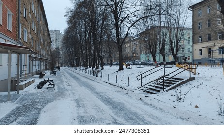 Concrete tile stairs with metal handrails lead from the asphalt road and apartment building up the slope to other buildings. There are trees, parked cars and everything is covered in snow. Overcast - Powered by Shutterstock