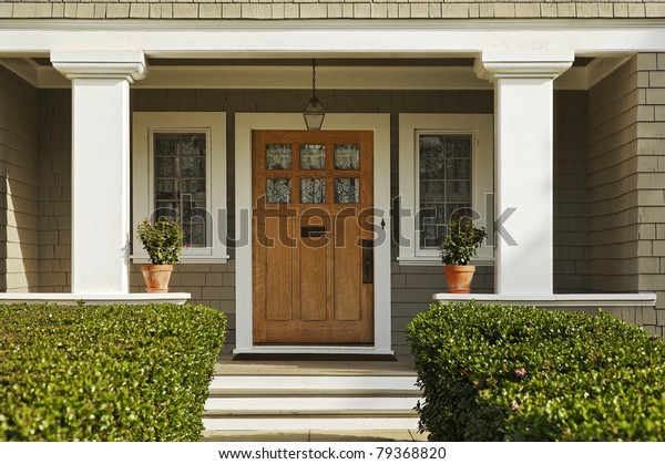 A concrete walkway bordered with hedged\
shrubs leads to the front door of a home. There are windows on\
either side of the door. Horizontal\
shot.