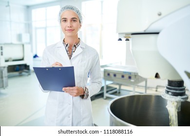 Confectionery Factory Employee Standing In White Coat Near Operating Machinery Holding Clipboard And Looking At Camera. 