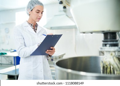 Confectionery Factory Employee Standing In White Coat Near Operating Machinery And Making Notes. 