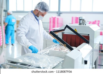 Confectionery factory employee in white coat wrapping paper box with pastry into packaging film.