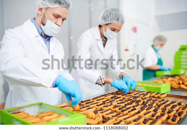 Confectionery factory workers\
in white coats making pastry with chocolate cream and putting it\
into boxes.