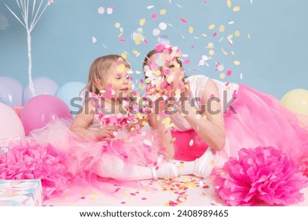 A confetti birthday party celebration. A mother and a little daughter are sitting in pink skirts on blue background. 