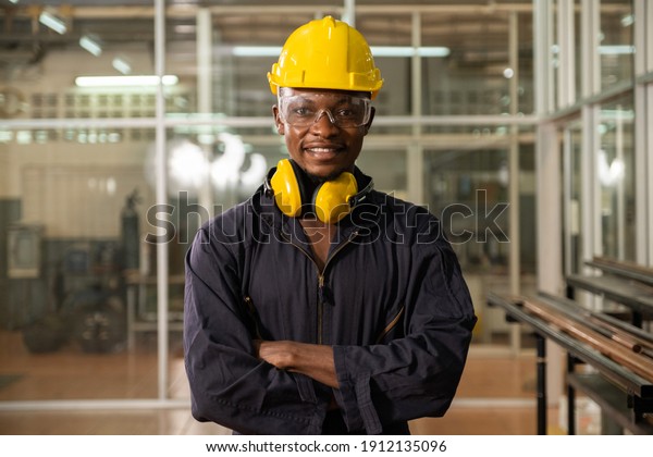 Confidence African American engineer or worker\
crossing his arms and smiling in the factory. Professional Heavy\
Industry engineer or worker Wearing Uniform, Glasses and Hard Hat\
in a Steel factory.