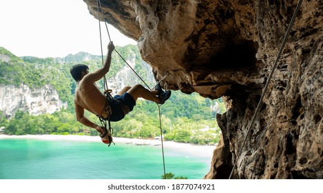 Confidence Asian Man Climber With Safety Rope Climbing High Up On Rocky Mountain At Tropical Island In Summer. Healthy And Strong Male Enjoy Active Lifestyle And Extreme Sport In Holiday Vacation.
