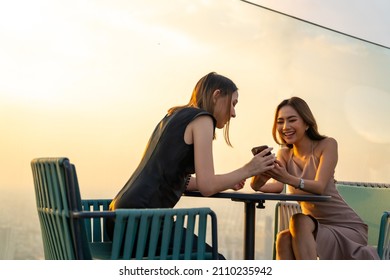 Confidence Asian Woman Friends Using Smartphone For Social Media Or Online Shopping Together While Meeting And Having Dinner At Skyscraper Outdoor Rooftop Restaurant Bar In The City At Summer Sunset