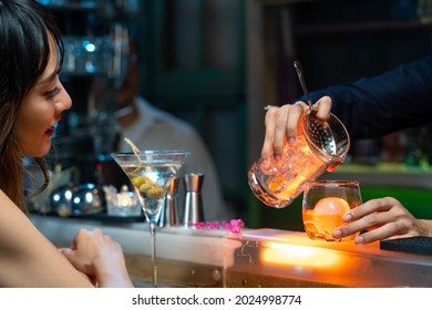 Confidence Asian Woman Sitting At Bar Counter Drinking Alcoholic Drink And Talking To Barman In Nightclub. Male Mixologist Bartender Preparing Alcohol Drink To Customer. Small Business Bar And City Ni