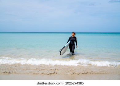 Confidence Asian Woman In Wetsuit Holding Surfboard And Walking On Tropical Beach At Summer Sunny Day. Healthy Sportswoman Enjoy Outdoor Active Lifestyle Play Extreme Sport Surfing On Summer Vacation