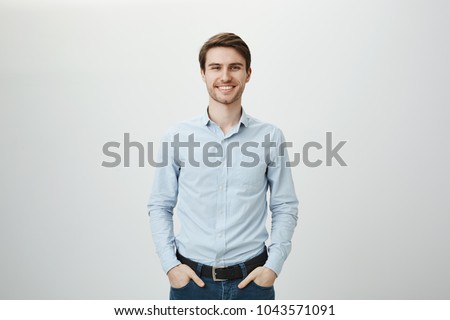 Confidence and business concept. Portrait of charming successful young entrepreneur in blue-collar shirt, smiling broadly with self-assured expression while holding hands in pockets over gray wall