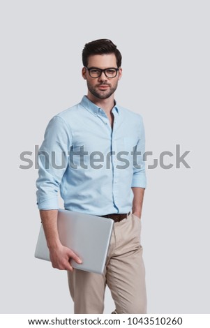 Confidence and charisma.  Good looking young man carrying laptop and looking at camera with smile while standing against grey background