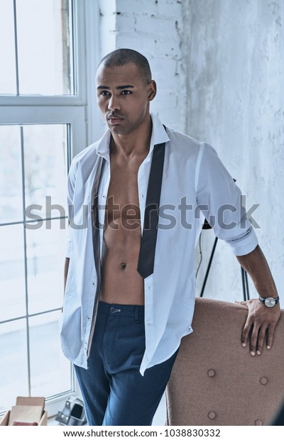 Confidence and\
charisma. Handsome young African man in fully unbuttoned white\
shirt looking away while standing at\
home
