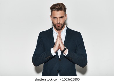 Confidence And Charisma. Handsome Young Man In Formalwear Holding Hands Clasped And Looking At Camera While Standing Against Grey Background