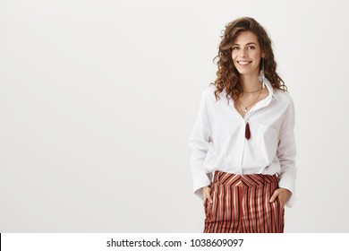Confidence is new sexy. Portrait of charming curly-haired woman in trendy garment smiling broadly and glancing at camera while discussing new plan for today with team over gray background.