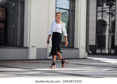 Confidence and sophistication mature woman walks city streets in fashionable ensemble chic white shirt and black fringed skirt with high heeled sandals and socks, fashion icon of style. - Powered by Shutterstock