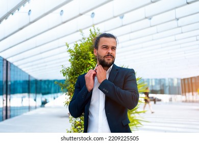 Confidence Successful Businessman Or Worker In Suit With Beard Standing In Front Of Office Building Straightening His Shirt. Hispanic Male Business Person Portrait Looking Away.