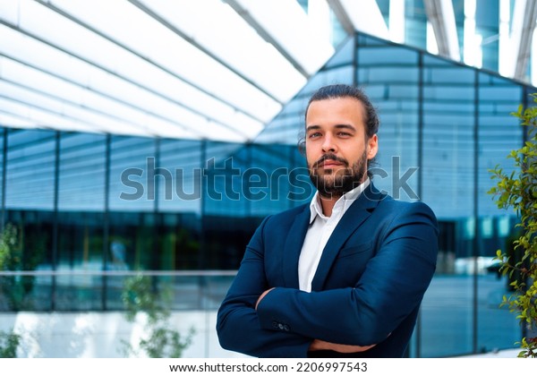 Confidence successful businessman or worker in\
suit with beard standing in front of office glass building arm\
crossed. Hispanic handsome business man looking at camera. Portrait\
shoot. Bank worker