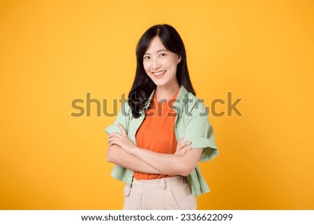 confidence and well-being with young 30s Asian woman wearing orange shirt portrays strength through arm cross gesture on chest against yellow background, a powerful portrait of self-assuredness.