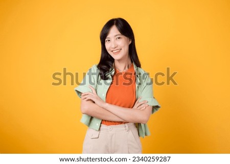 confidence and well-being with young 30s Asian woman, wearing orange shirt. Her arm cross gesture on her chest against a vibrant yellow background paints portrait of self-assuredness and inner peace.