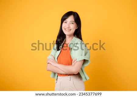 confidence and well-being with a young 30s Asian woman wearing orange shirt. Her arm cross gesture on chest against a vibrant yellow background paints a portrait of self-assuredness and inner peace.