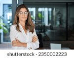 Confident 45 year old Latin professional mid aged business woman corporate leader, mature female executive manager wearing eyeglasses standing in office arms crossed looking at camera, portrait.