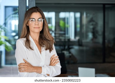 Confident 45 year old Latin professional mid aged business woman corporate leader, mature female executive manager wearing eyeglasses standing in office arms crossed looking at camera, portrait. - Powered by Shutterstock