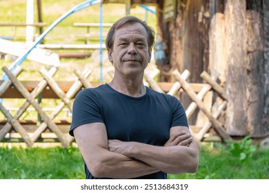 A confident 50-55 year old elderly man stands with arms crossed in casual outdoor clothing near a wooden structure. - Powered by Shutterstock