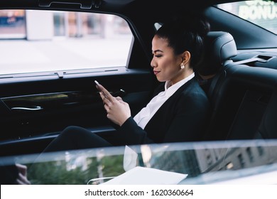 Confident Adult Asian Executive Woman In Formal Clothes Concentrating On Screen And Interacting With Smartphone While Sitting In Luxurious Automobile In City
