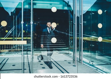 Confident Adult Businessman In Black Suit Leaving Office Building Walking Through Glass Doors In Business Center In City In Summer Day