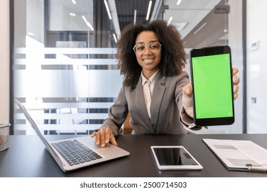 Confident African American businesswoman sitting at desk showing phone with green screen. Professional attire, using laptop and tablet, modern workspace, technology, and business concepts - Powered by Shutterstock